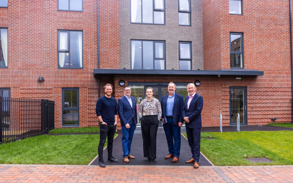 Men and women representing the delivery partners stand in front of a red and grey brick-fronted building to mark the completion of Trafford Waters Care Home.